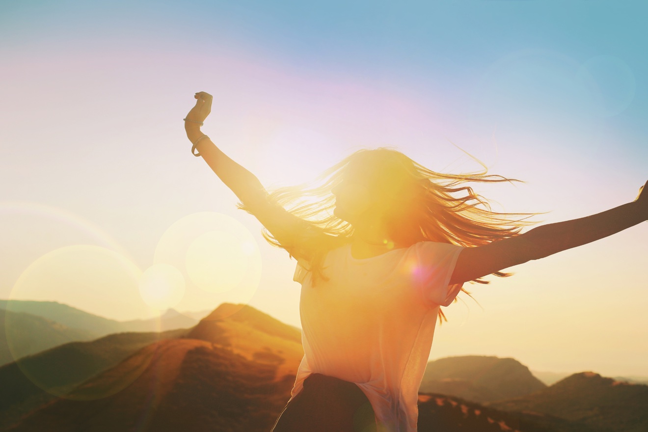 A woman with her arms raised wide at sunset, backlit by golden light over mountain ridges